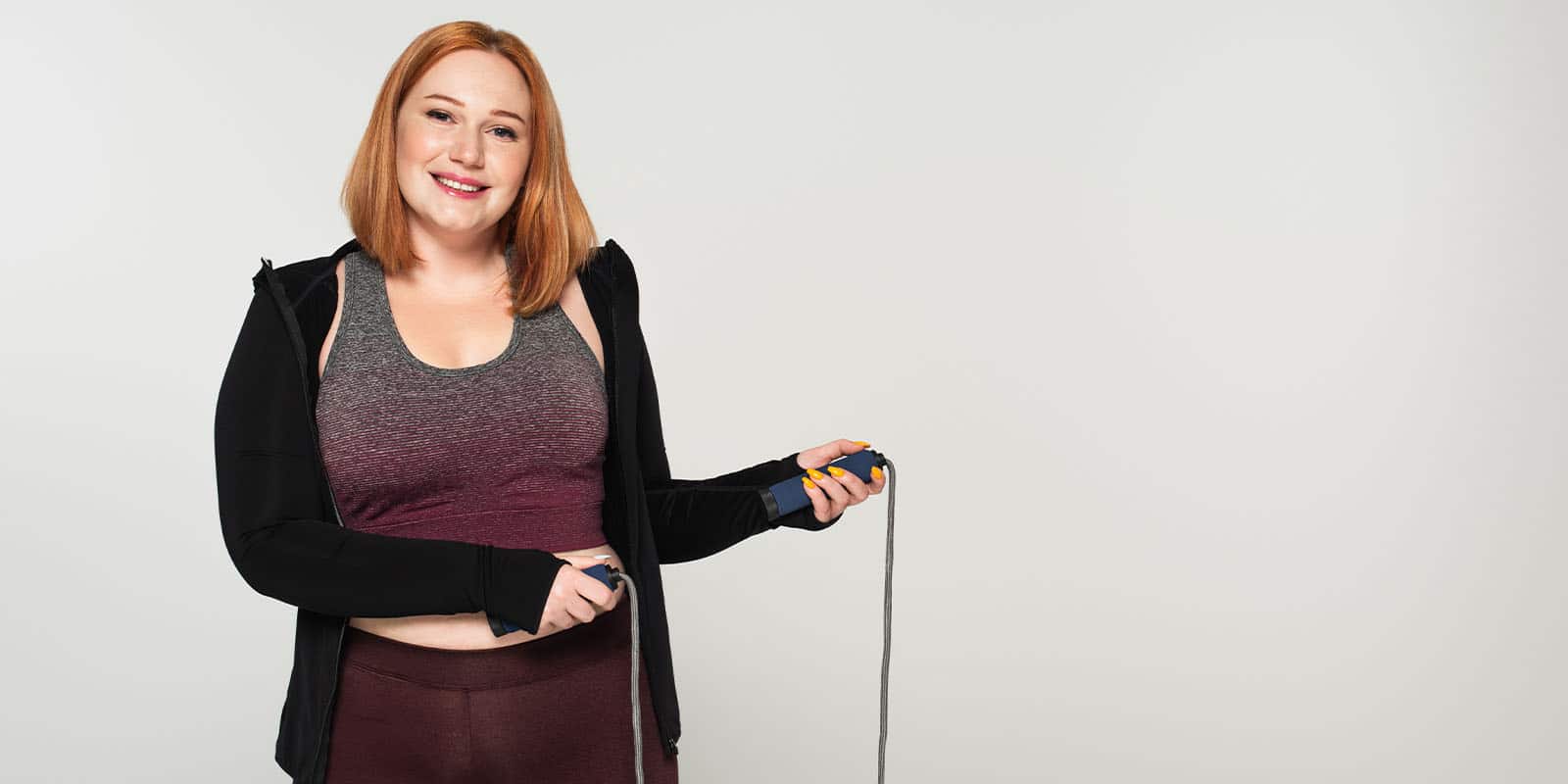 A body-positive woman in athletic wear holding a jump rope and smiling against a plain white background.