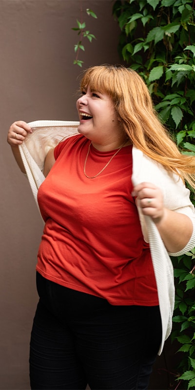 A cheerful plus-size woman with red hair, wearing an orange shirt and white cardigan, laughing and posing in front of green vines.