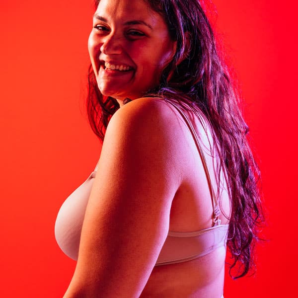 A smiling plus-size woman with long dark hair, wearing light-colored lingerie, posing in a studio with a red background.