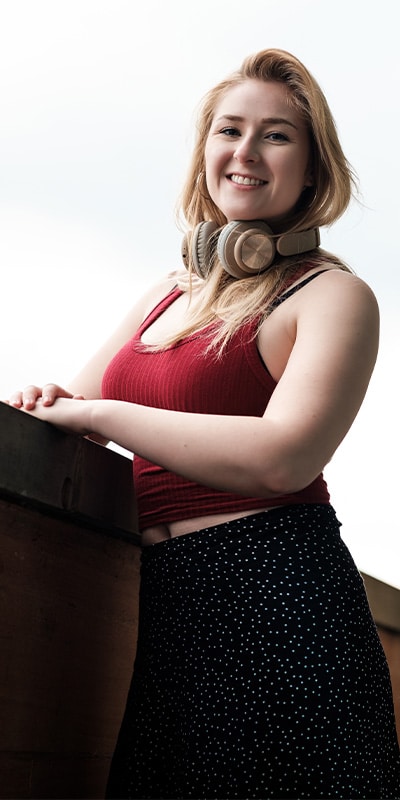 A young woman in a red tank top with headphones around her neck, leaning against a wooden railing and smiling.